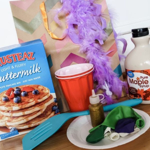 Box of pancake mix, red plastic cups, a blue plastic spatula, a bottle of maple syrup, a gift bag with a purple feather boa, and a white paper plate with uninflated balloons in various colors