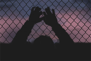 A person grasping a chain-linked fence is silhouetted against a sky that is almost dark.