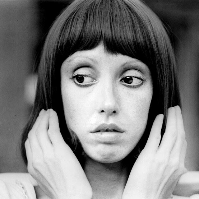 Shelley Duvall in a black-and-white portrait from the 1970s, holding her hands up to the sides of her head.