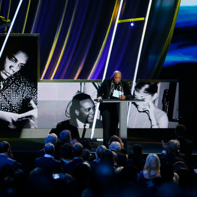 Quincy Jones, wearing a black suit, stands on stage in front of screens showing images of him. 