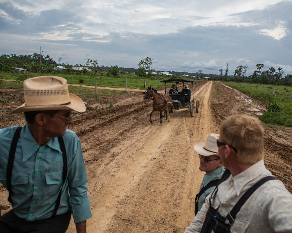 Two people ride in a horse and buggy on a dirt road as three men watch.