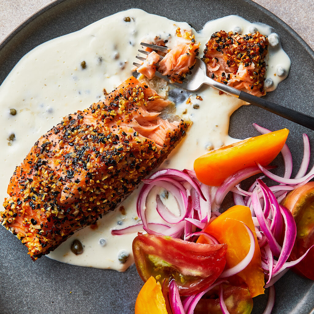 A plate of salmon, topped with seasoning, sitting on a cream sauce and accompanied by red onion and tomatoes. 