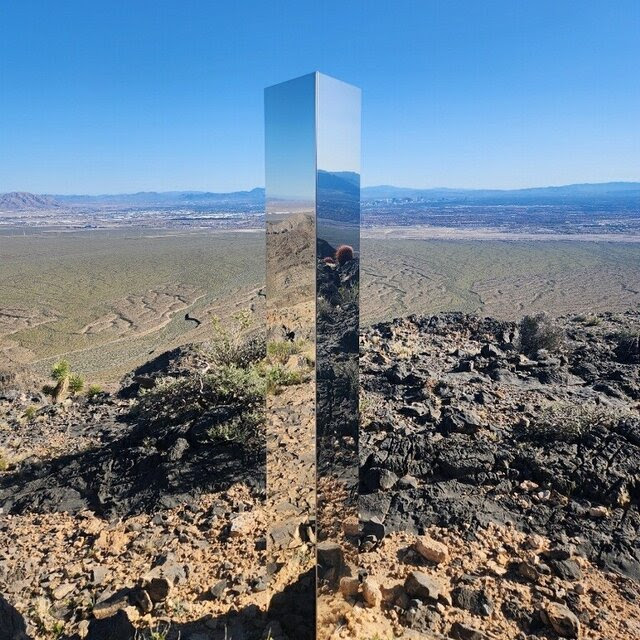 A slick rectangular column mirrors the rough terrain and sky in desert land.