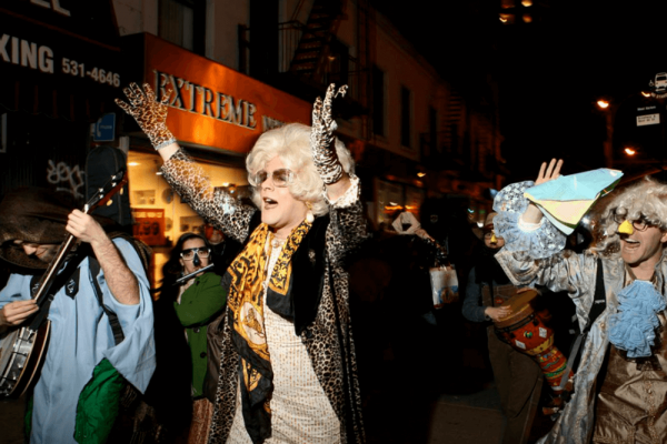 Drag queen wearing voluminous platinum blond wig, a leopard print silk cardigan, leopard print gloves, and a gold and black scarf puts her arms in the air amidst a crowd.