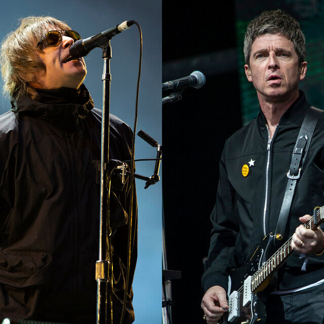 A man in shaggy hair, sunglasses and a zipped up windbreaker sings into a microphone; a man in a zipped up black jacket plays guitar.