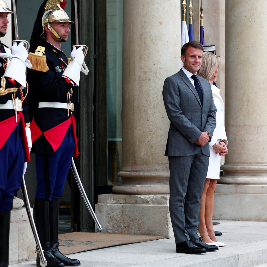 A pair of soldiers in ceremonial uniform stand to the side of President Emmanuel Macron of France and his wife.