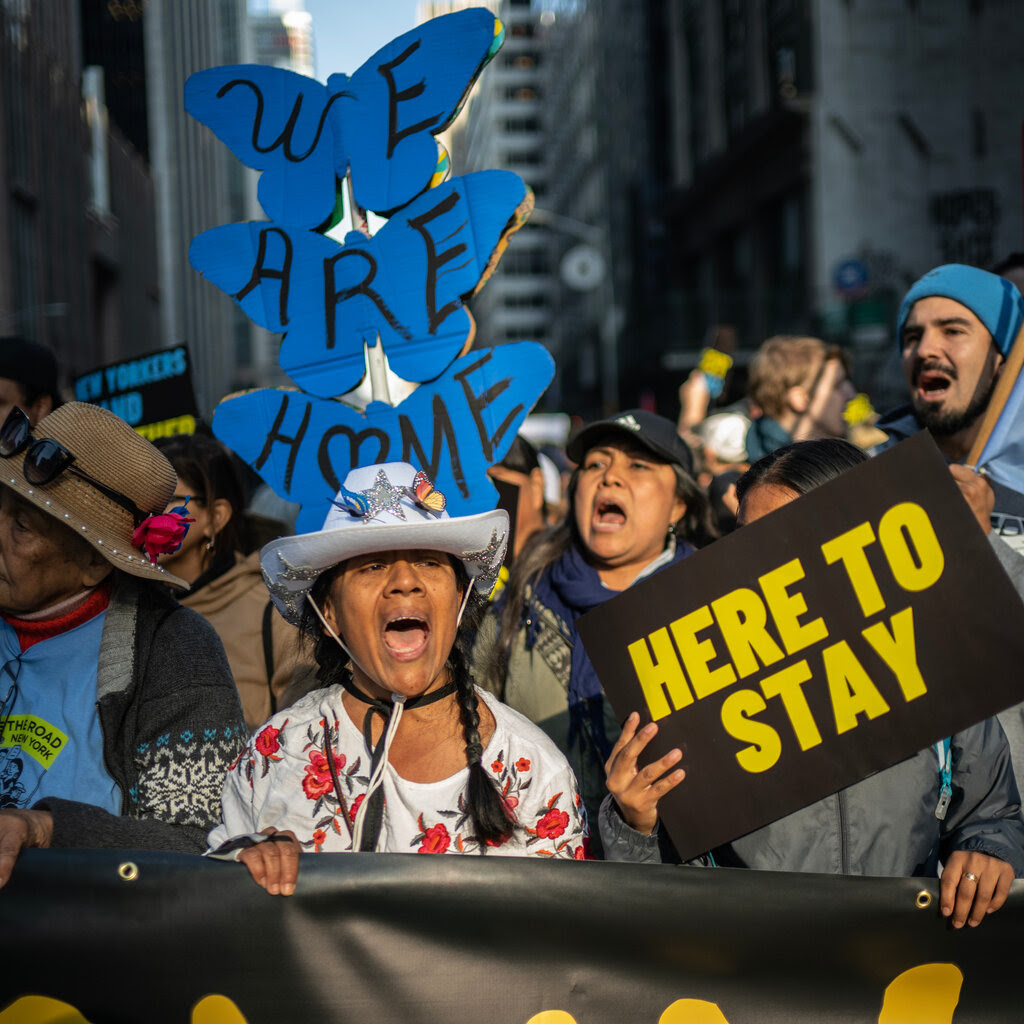 Several people marching in a protest as they hold a banner. One person holds up a sign that says “Here to Stay.” Another sign reads “We Are Home.” 