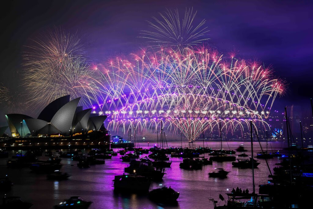 Fireworks light up the sky over a lit-up Harbour Bridge in Sydney, Australia. Boats float on the harbor in the foreground.