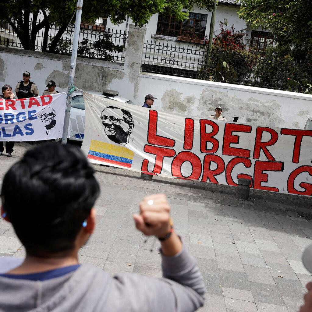 People holding a banner reading “Libertad Jorge Glas” near a cement wall. A person with a raised fist and facing away from the camera is in the foreground.