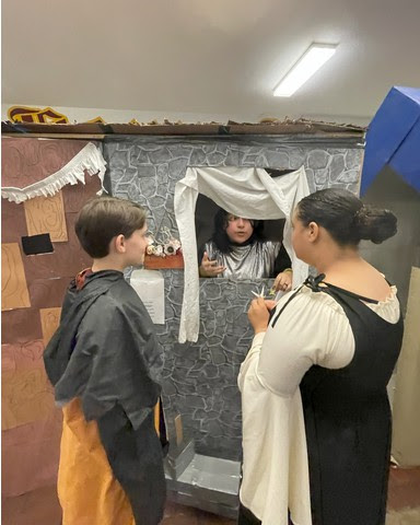 Two students dressed in medieval clothing speak to another student through a window, they are inside a cardboard structure painted to look like a stone building 