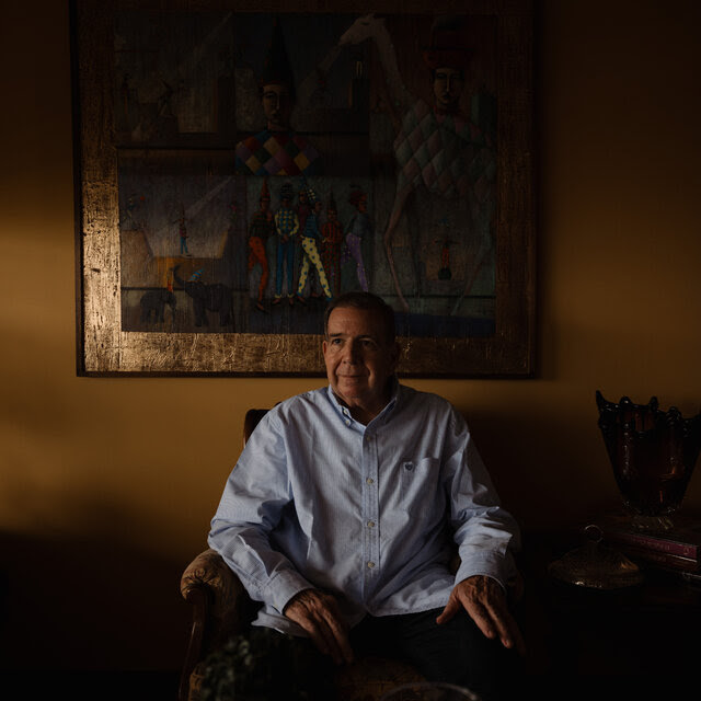 A man in a blue shirt sits in front of a painting in a room in shadow. 