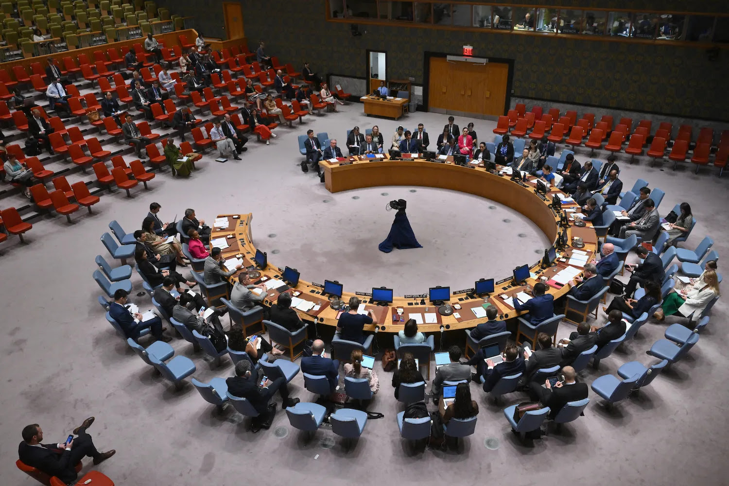 An overheard view shows the large central room at the U.N. headquarters. Representatives of members of the Security Council sit at a large horseshoe-shaped desk at center as they attend a meeting.