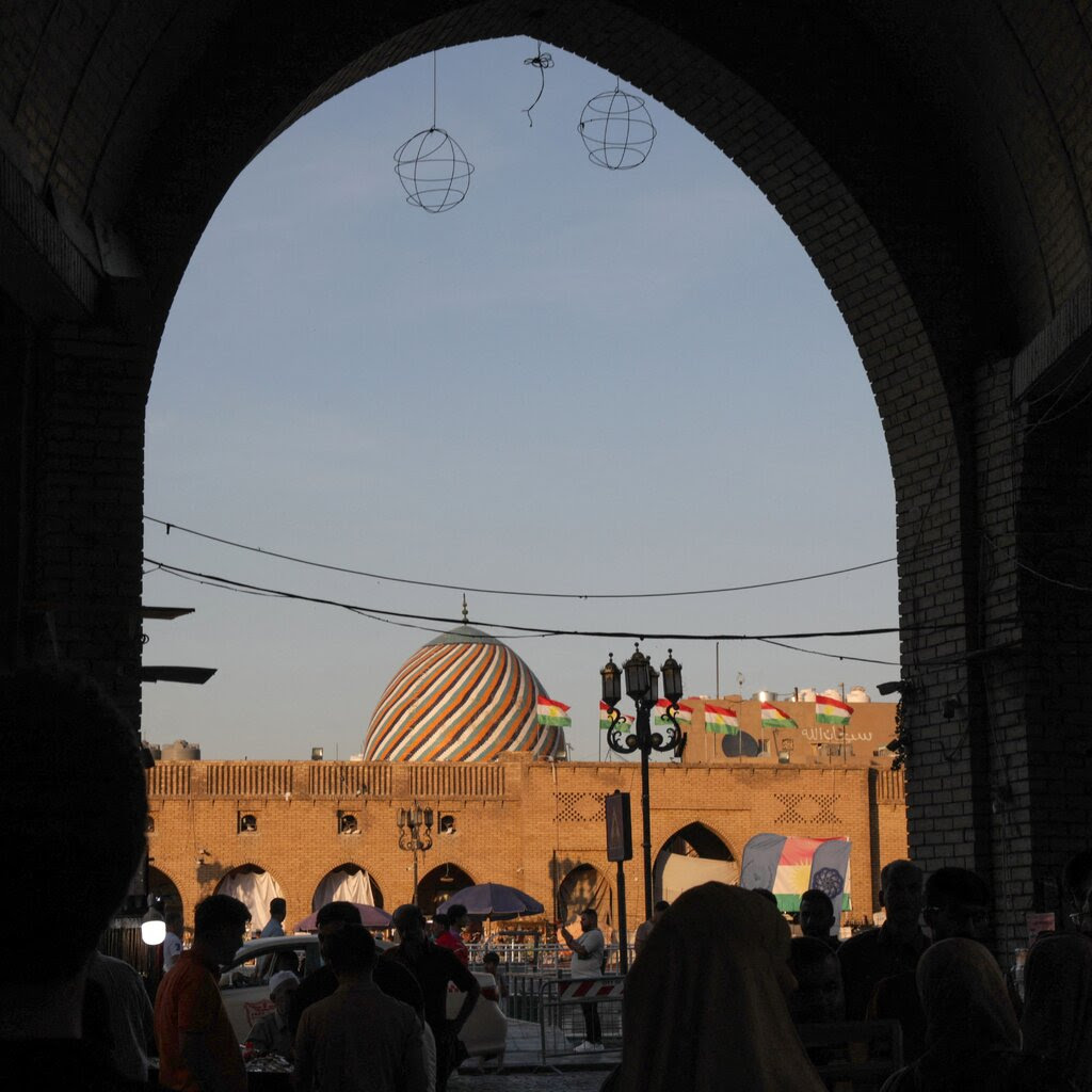 People milling around outdoors. A striped dome rises over a brick wall in the background.