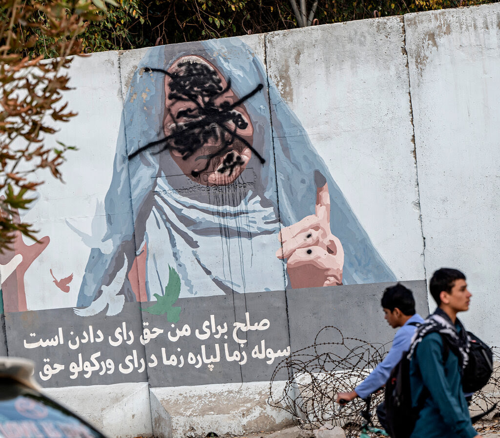 Young men ride or walk past an outdoor mural near a vehicle and tree in daylight.