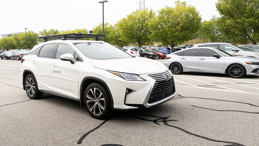 A white, Purdue-branded midsize crossover vehicle drives through a parking lot