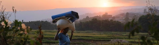 photo of woman carrying fabric and bag on her head in an open field