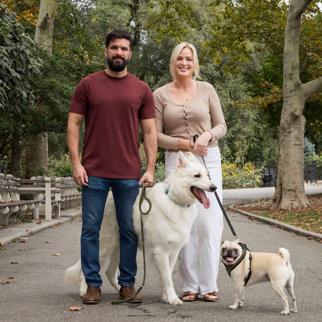 A man in a maroon T-shirt and a woman in a tan top stand in a city park, each of them holding a dog on a leash.