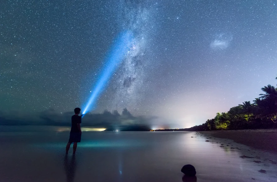 A person shines a flashlight into the sky while standing on a beach at night