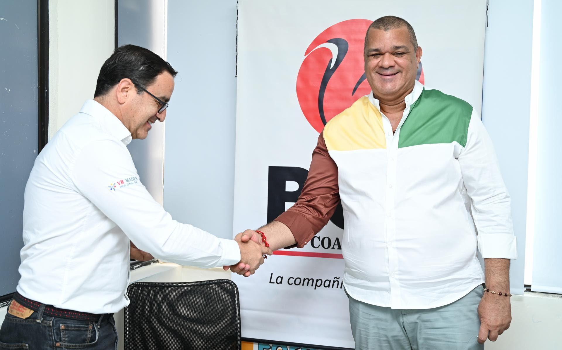 Dos hombres de negocios dándose la mano. Uno de ellos, vestido con una camisa blanca con detalles de colores verde, amarillo y marrón, sonríe. El otro, con una camisa blanca y gafas, lleva un micrófono de solapa. Detrás de ellos hay un logotipo corporativo de la compañía y el nombre de una universidad.