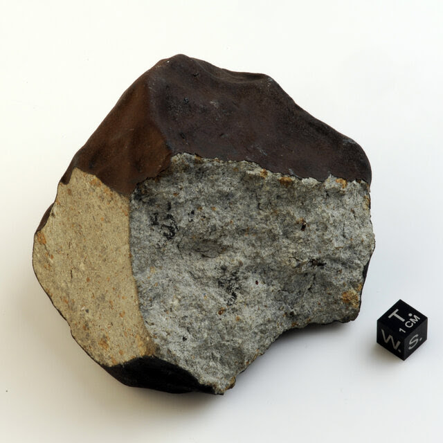 A close-up view of a meteorite chunk on a white surface with two sides that are a lighter color than its dark black top.