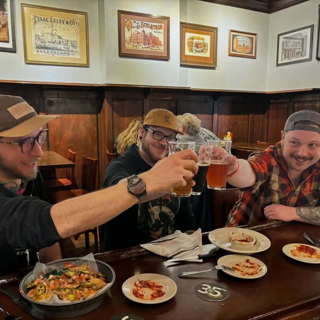 Three GLBC employees toasting with GLBC beers of various hues, with all day happy hour appetizers on the bar in front of them.