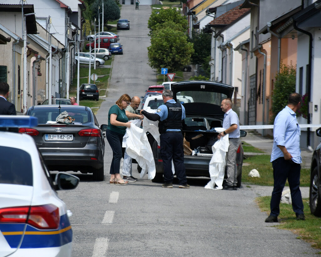 People in plain and police clothing standing behind a car with an open trunk. They hold large pieces of white cloth on a residential street with a police vehicle in the foreground.