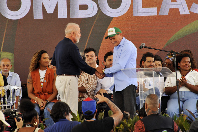 Em palco de evento, presidente Lula aperta mão de homem idoso, usando camisa branca