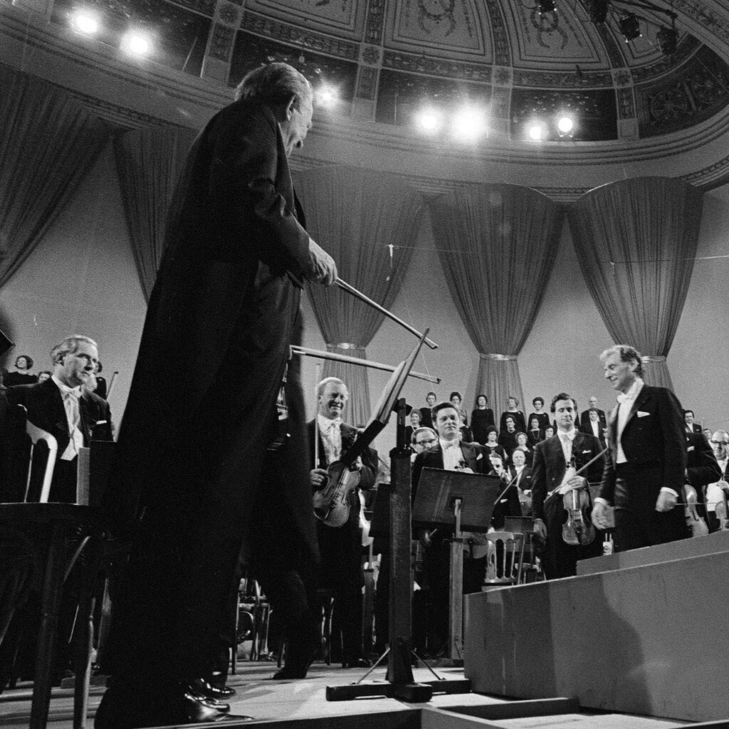 A black-and-white historical photograph shows Leonard Bernstein onstage in a concert hall, surrounded by musicians in formal wear.