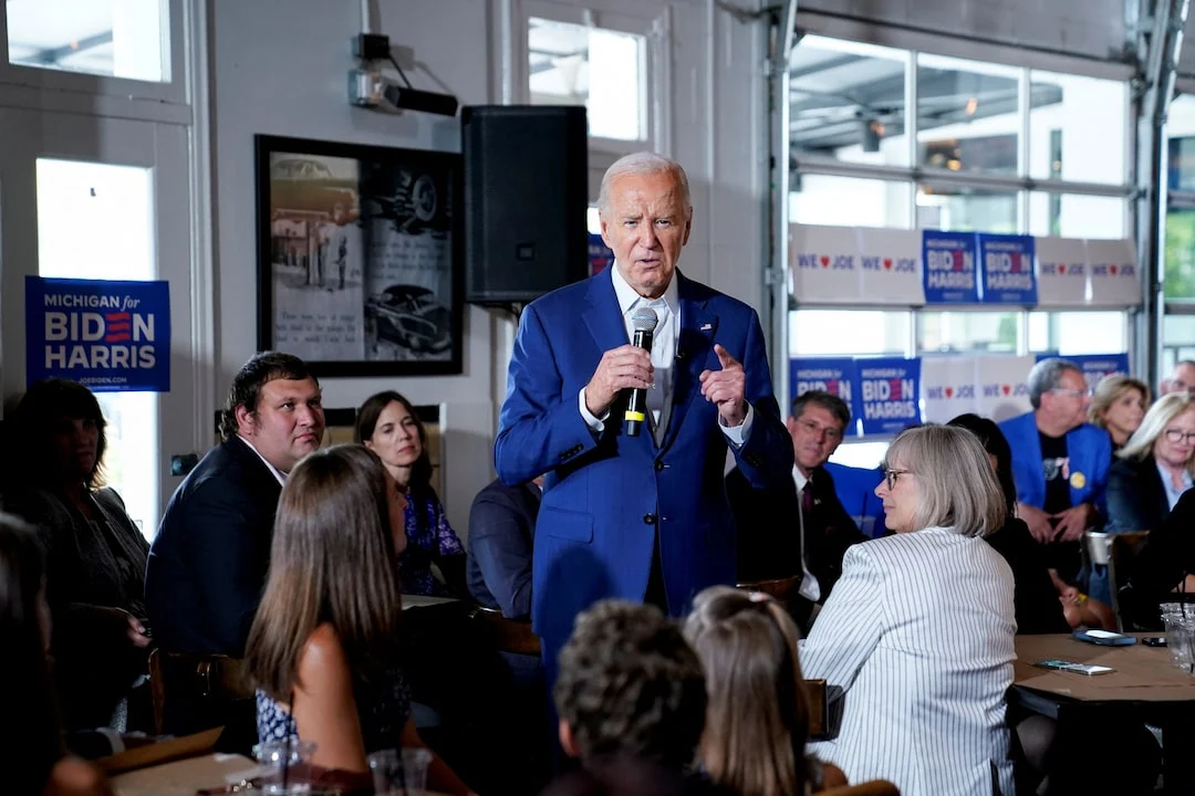 Joe Biden stands up holding a microphone as seated supporters at tables listen