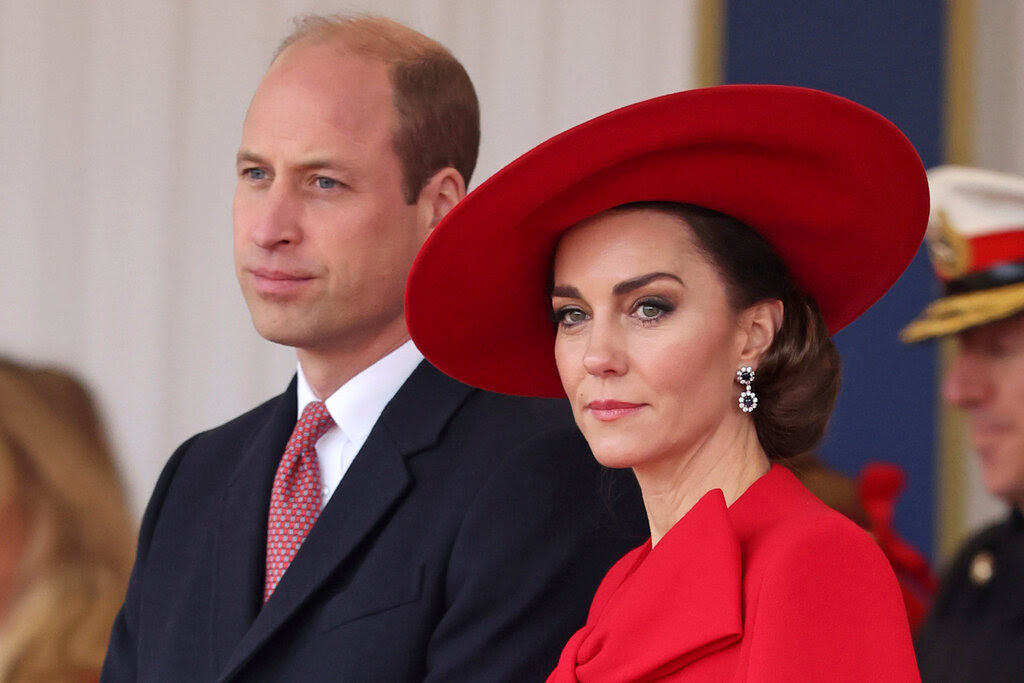 Prince William and Catherine, Princess of Wales, stand together. He wears a dark suit with a white shirt and red tie, while she wears a red outfit with a red hat.