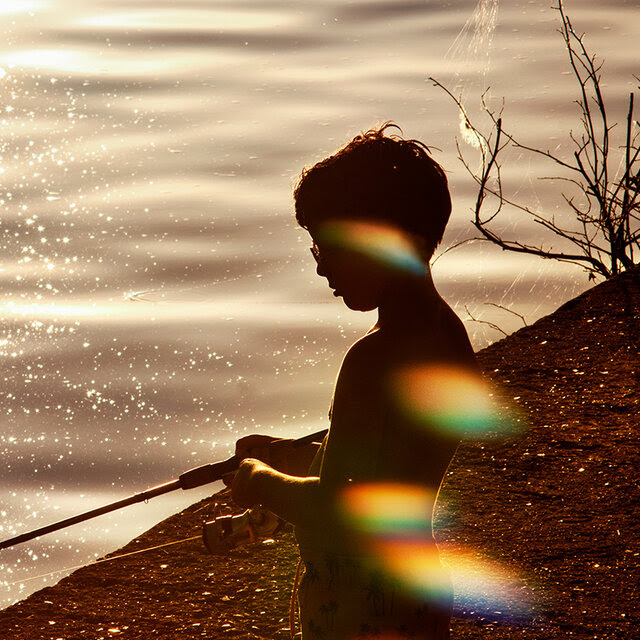 A boy in silhouette fishing on the shore of a lake. 