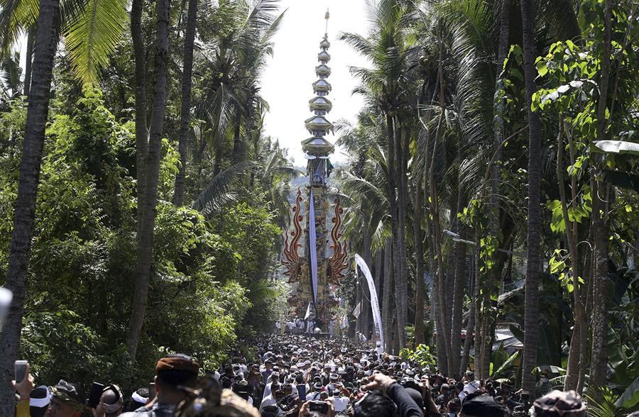 Men move a cremation tower in Indonesia. There is a procession of people following the tower and trees line either side of the pathway.