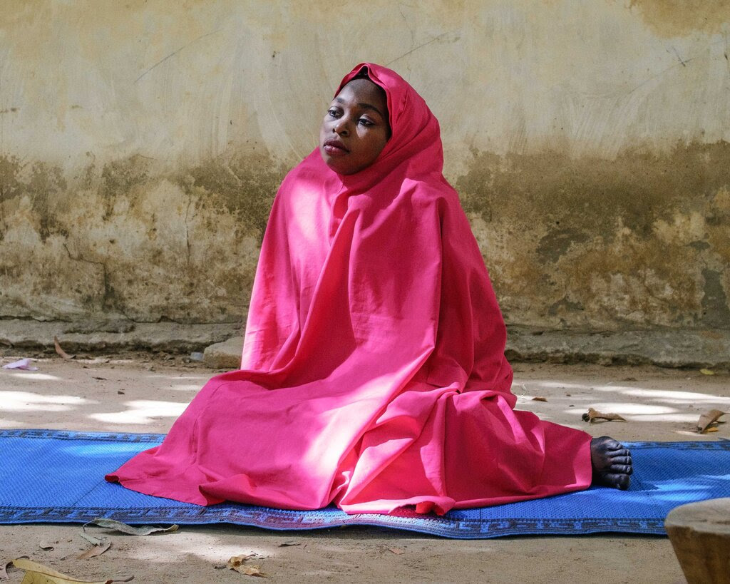 Saratu Dauda, in a bright pink head scarf and abaya, sits on a blue rug in front of an off-white building.