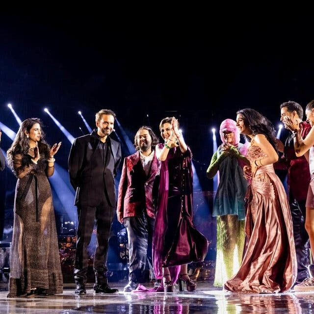 Nine people in fancy clothing stand on a stage, clapping, while spotlights shine down.