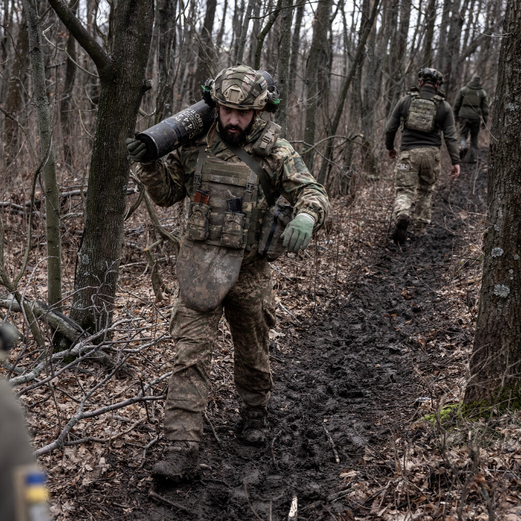 A bearded soldier in camouflage gear moves shell casings in the woods, as other soldiers share his path. 