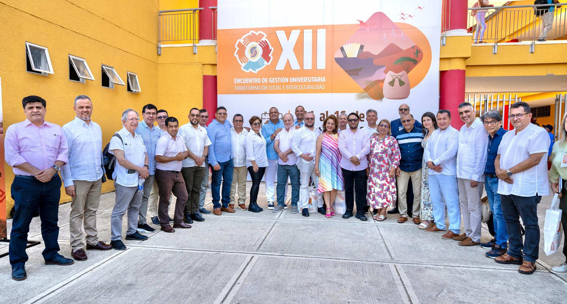 grupo grande de personas posando frente a un fondo que tiene el logo y texto del ''XII Encuentro de Gestión Universitaria''. Las personas están organizadas en dos filas frente a una pared amarilla, y varias de ellas están vestidas de manera formal, principalmente con camisas blancas. El grupo incluye tanto hombres como mujeres, y algunos de los participantes están sonriendo para la foto. La atmósfera parece ser la de un evento académico o institucional.