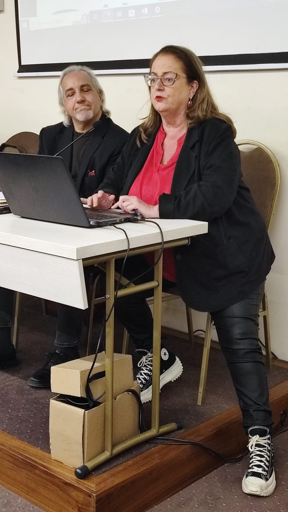 Smashing Times Company Manager Freda Manweiler, wearing blue jeans and a black jacket, making a presentation alongside a grey-haired gentleman