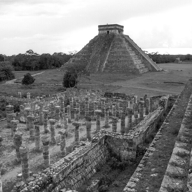 A black-and-white photo of the ruins of the ancient city of Chichén Itzá, including its iconic pyramid.