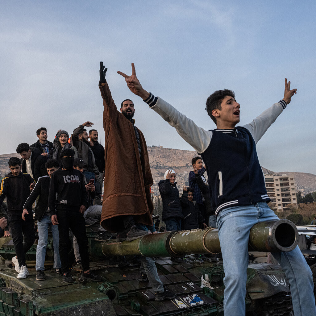 Men in street clothes on top of a tank, most of them smiling, a few waving, one of them straddling the barrel.