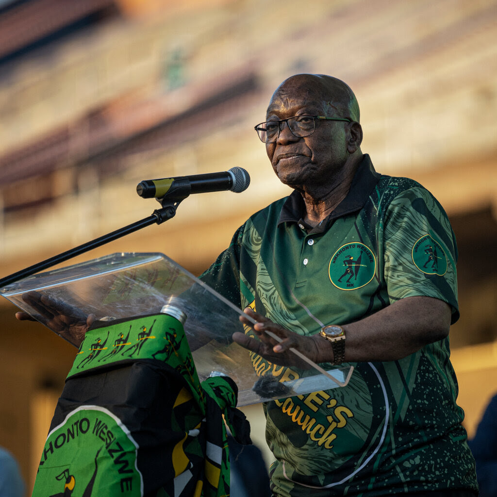 Jacob Zuma stands at a podium, wearing a green polo shirt.