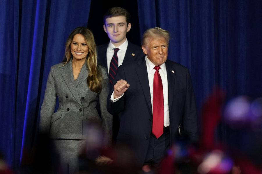 A light-haired man in a black suit and red tie, flanked by a smiling woman in a gray jacket and a tall young man, raises his fist.