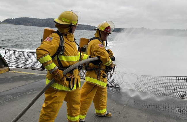 Two ferry crew members in firefighting gear on the car deck spraying water off the vessel