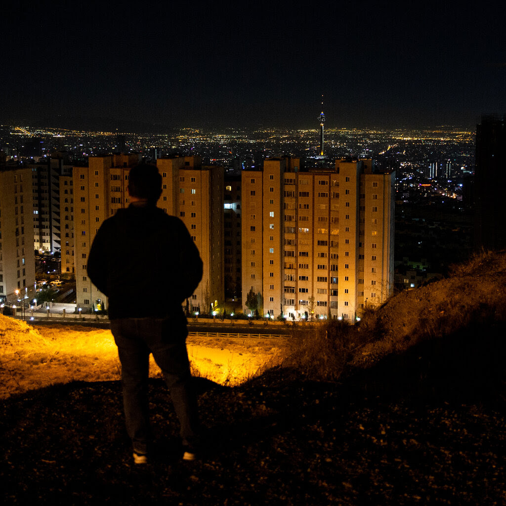 A person stands on a hill looking out at Tehran lit up at night.