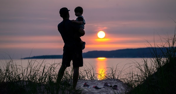 man in a ballcap holding a young boy, framed in shadow on the shore as the pink-orange sun sets on Little Traverse Bay