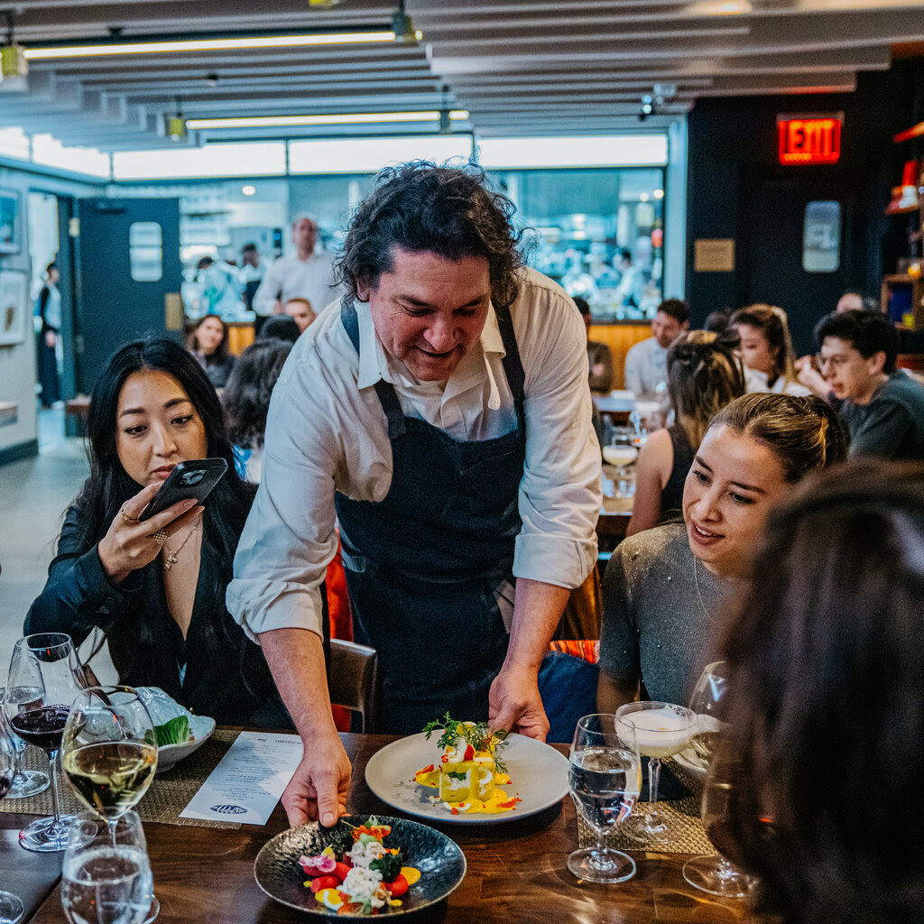 Chef Gaston Acurio shows off a dish to a table of smiling diners.