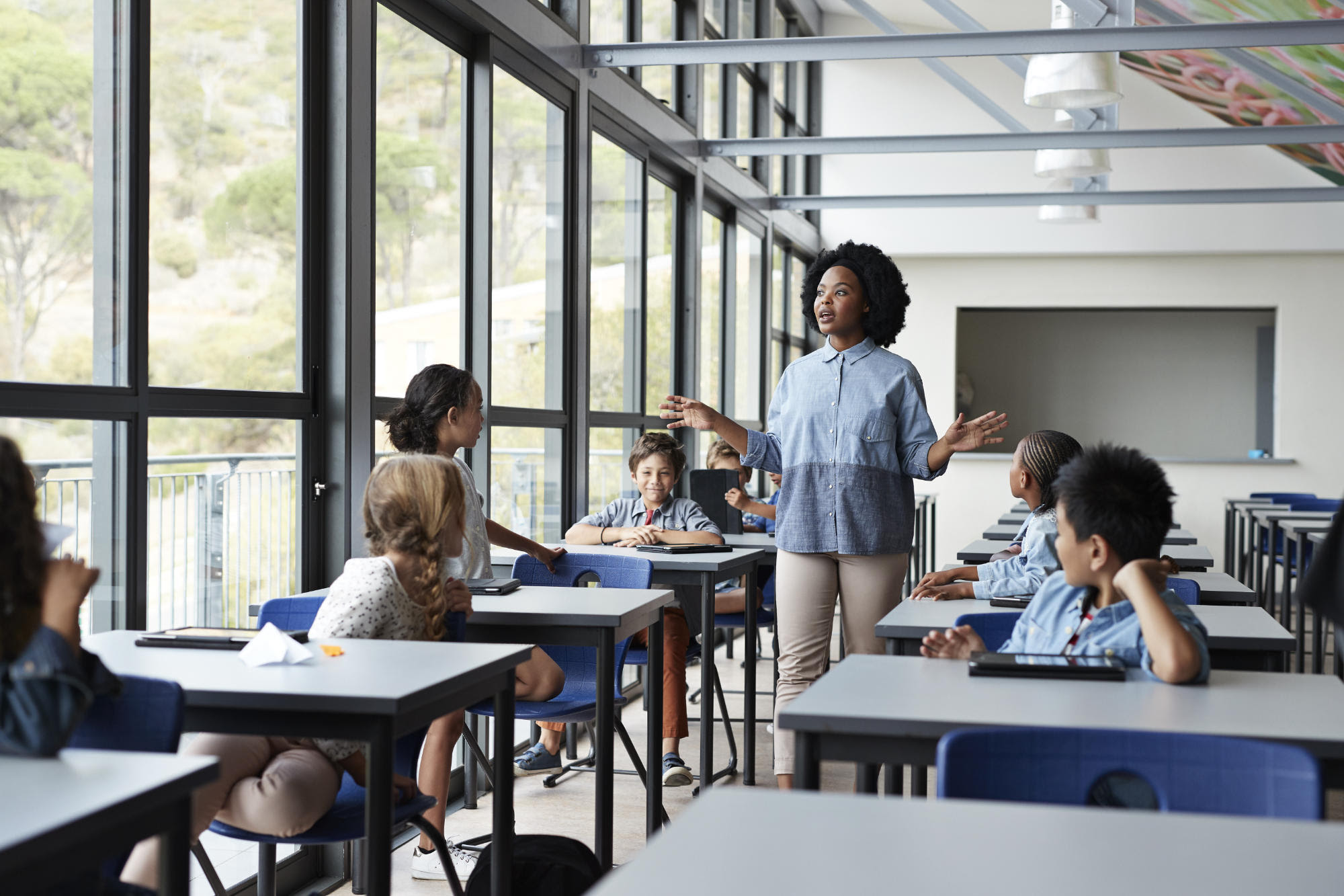 A group of children sitting at desks watch a teacher in a modern-looking classroom.