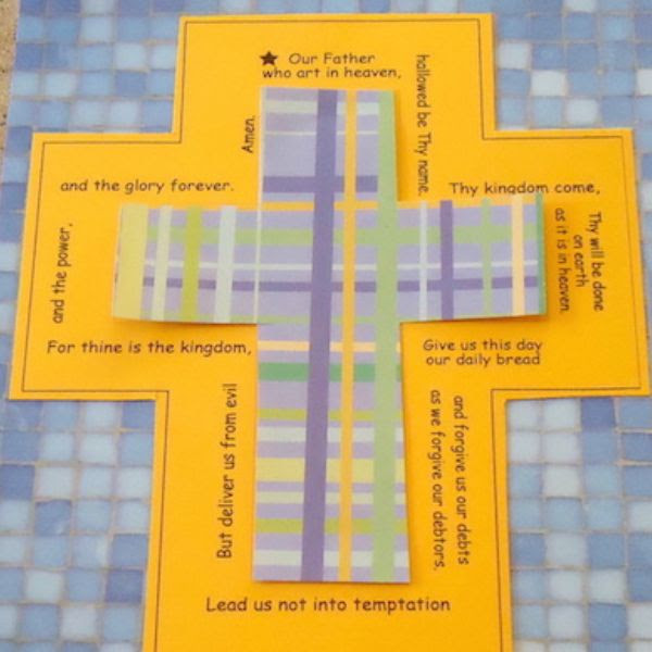 Blue, green, and yellow plaid paper cross on top of a yellow paper cross with the words of the Lord's Prayer on a blue tile background