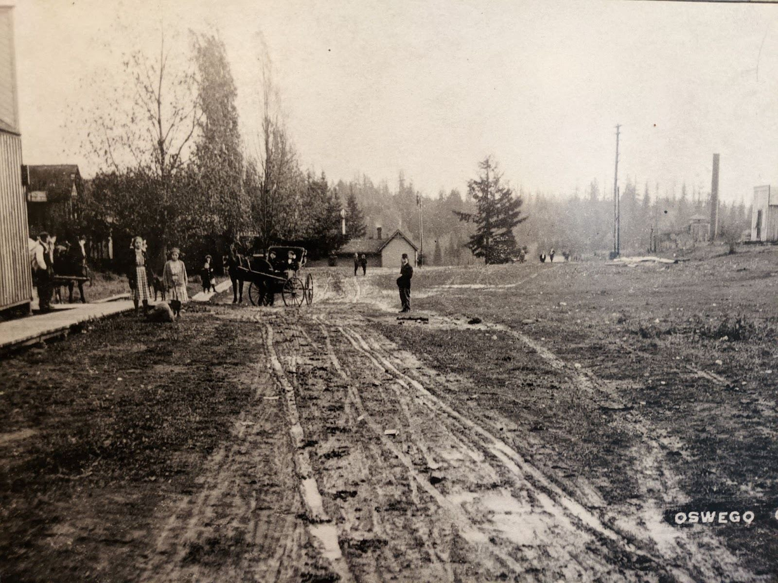 A circa 1890 street in Lake Oswego. The street is mostly mud with tracks on it. There is a coach being pulled by a horse and several people around.
