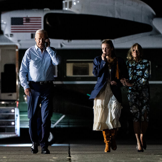 President Biden, wearing a blue shirt and blue pants, talks on a phone as he walks from a helicopter. He is flanked by family members, including Jill Biden, the first lady.