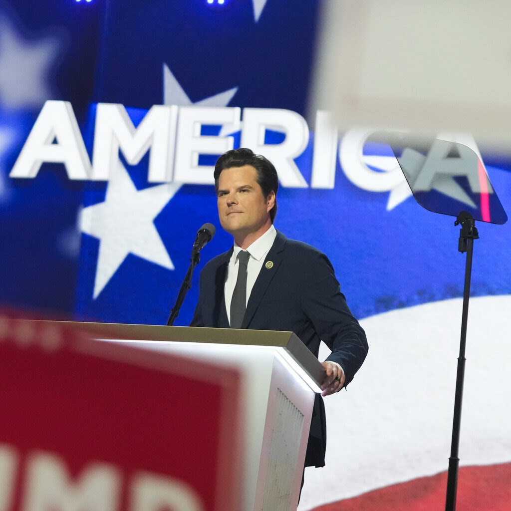 Matt Gaetz stands at a dais on the convention stage, with the word America on a screen behind him.